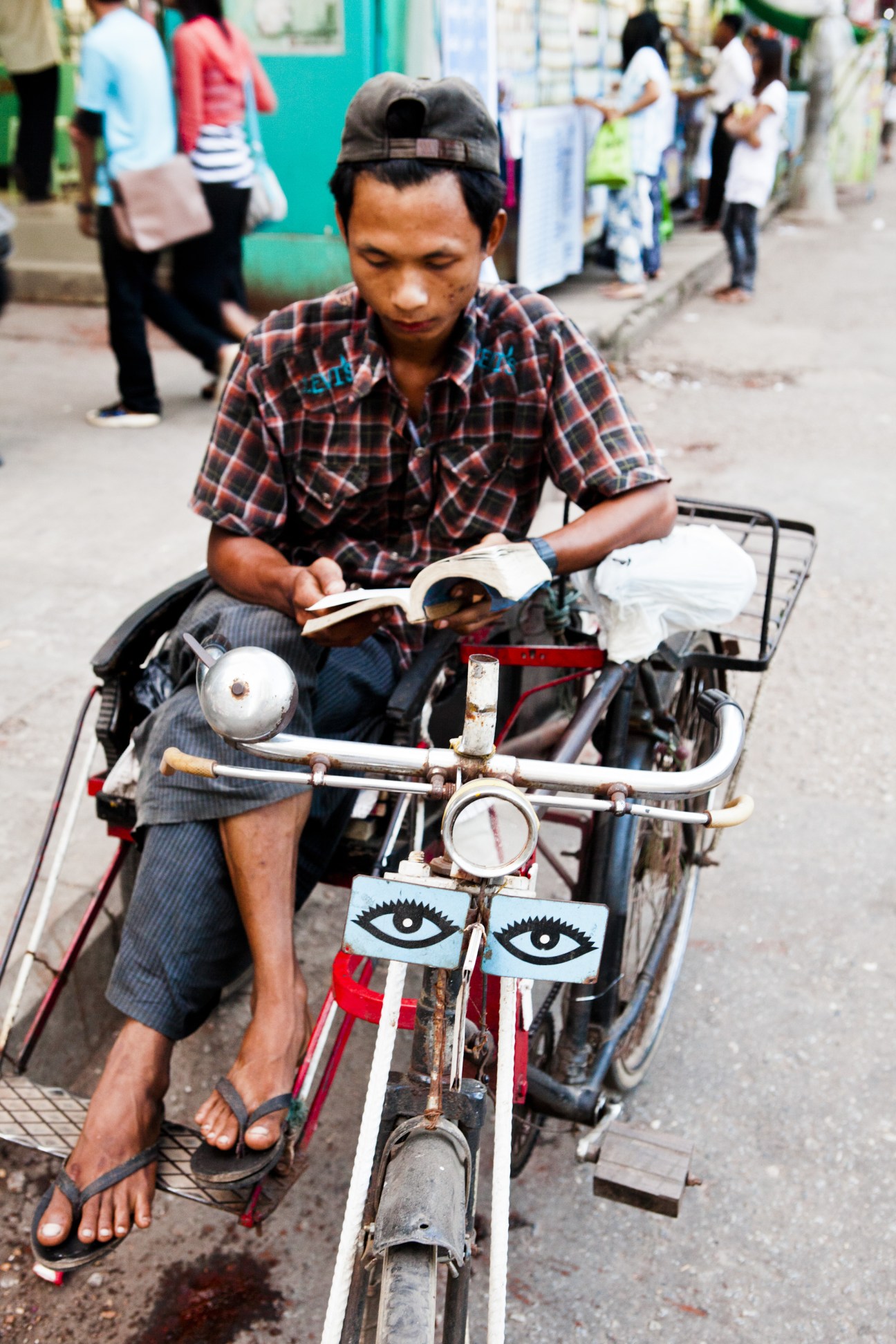 A trishaw driver reads a book while sitting on his decorated three-wheeled vehicle on a Rangoon street.