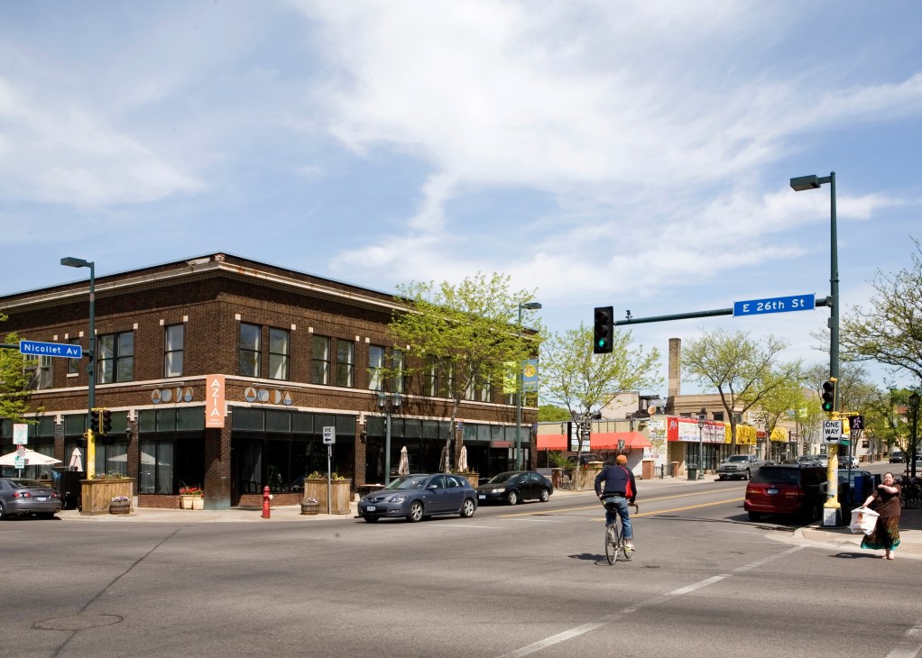 Intersection of Nicollet Avenue and E 26th Street in Minneapolis showing a brick corner building with ground-floor retail and restaurants.