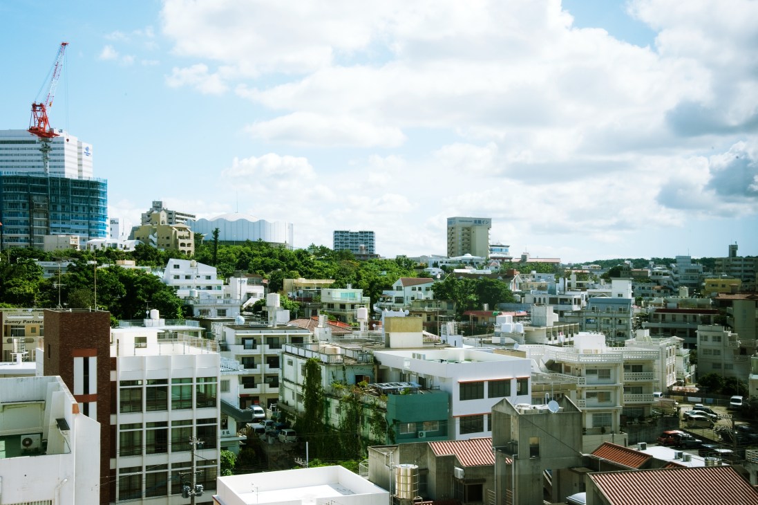 Aerial view of Naha cityscape with modern buildings, construction crane, and green hills under blue sky.