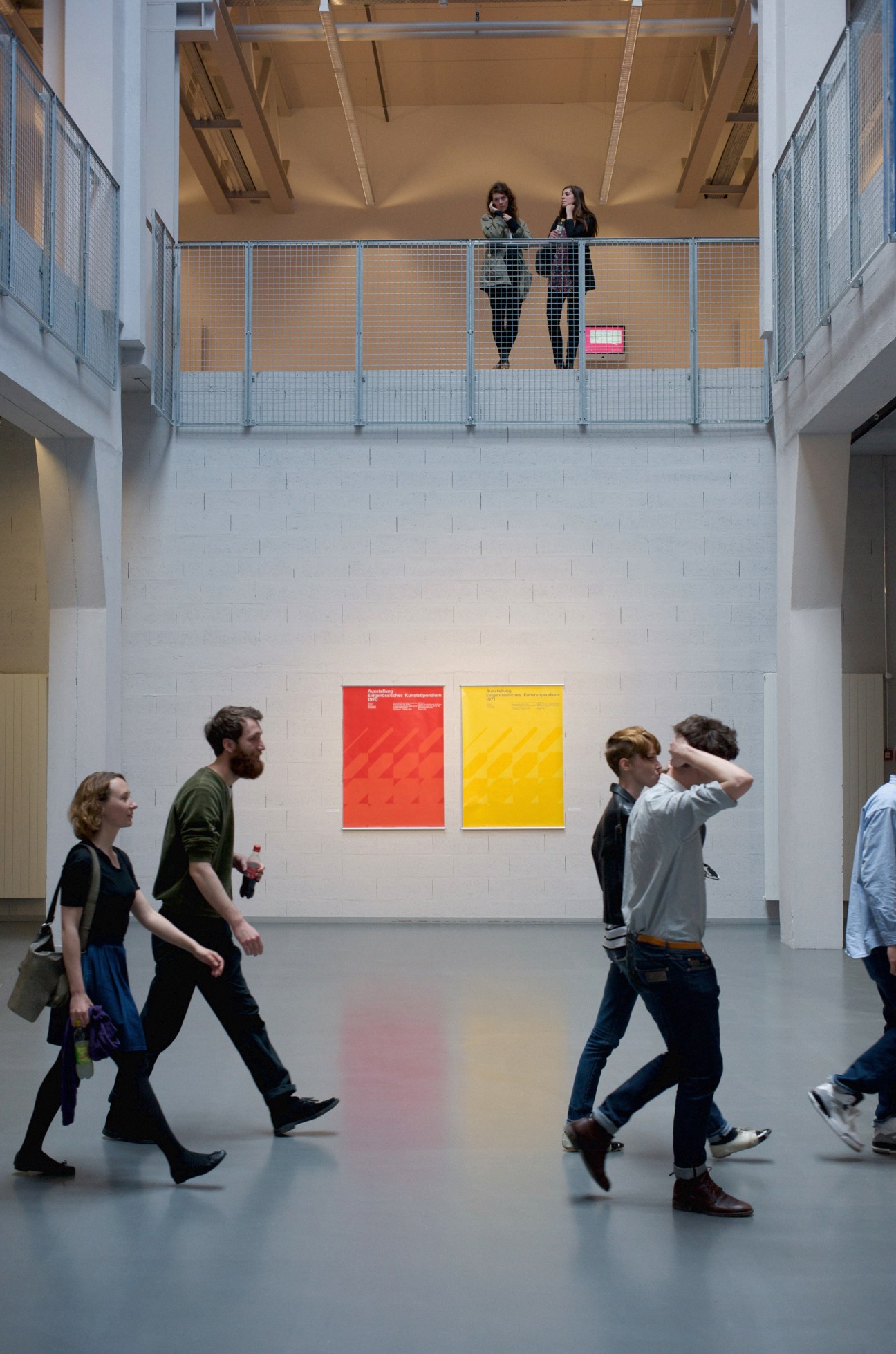 Students walking through a bright, minimalist gallery space with red and yellow posters on white walls.