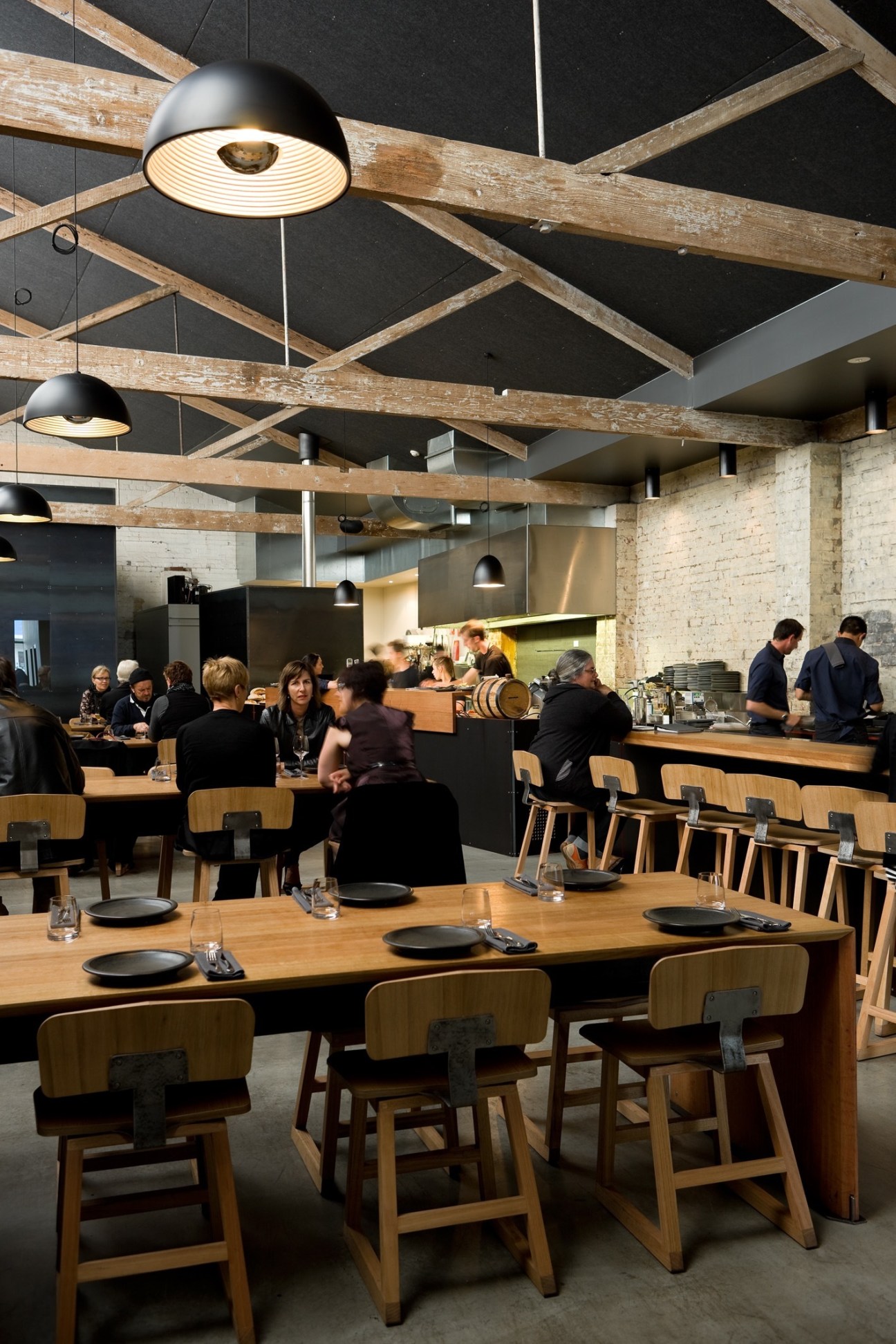 Modern restaurant dining room with exposed wooden beams, black pendant lights, and diners seated at wooden tables.