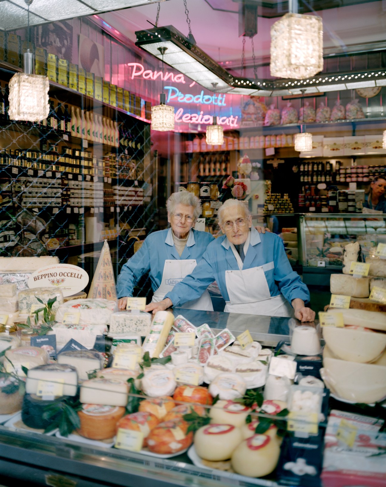 Two elderly women in blue uniforms stand behind a deli counter displaying various cheeses and cured meats at Latteria Bera Bruna.