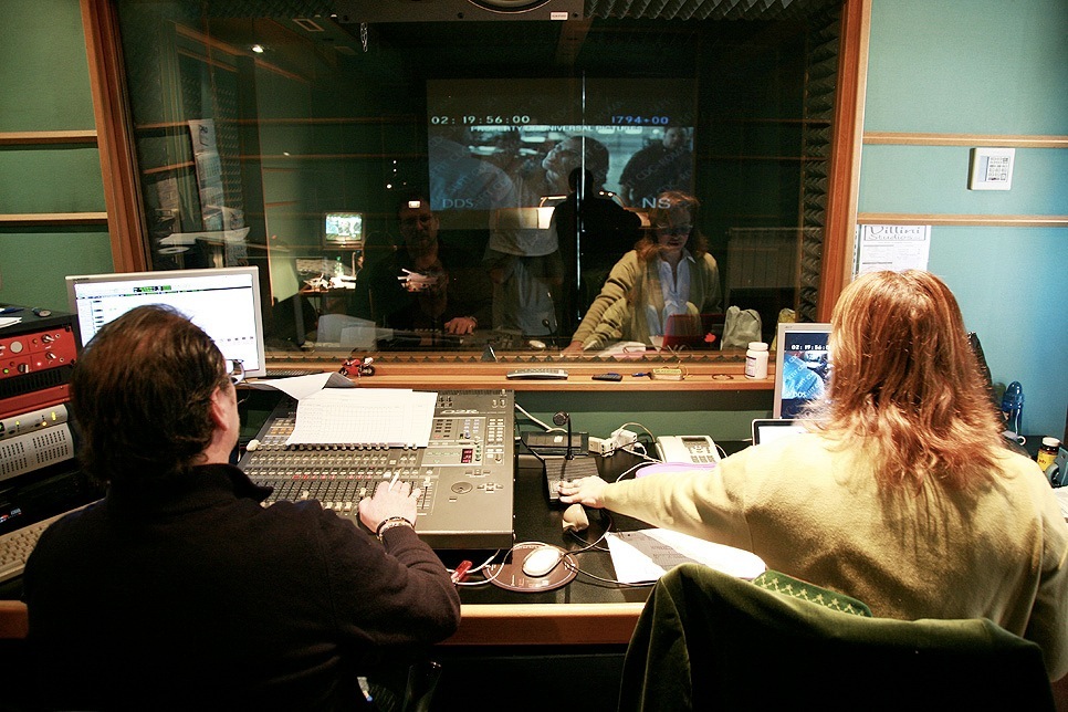 Voice actors recording dialogue in a dubbing studio with a director supervising through a glass window.