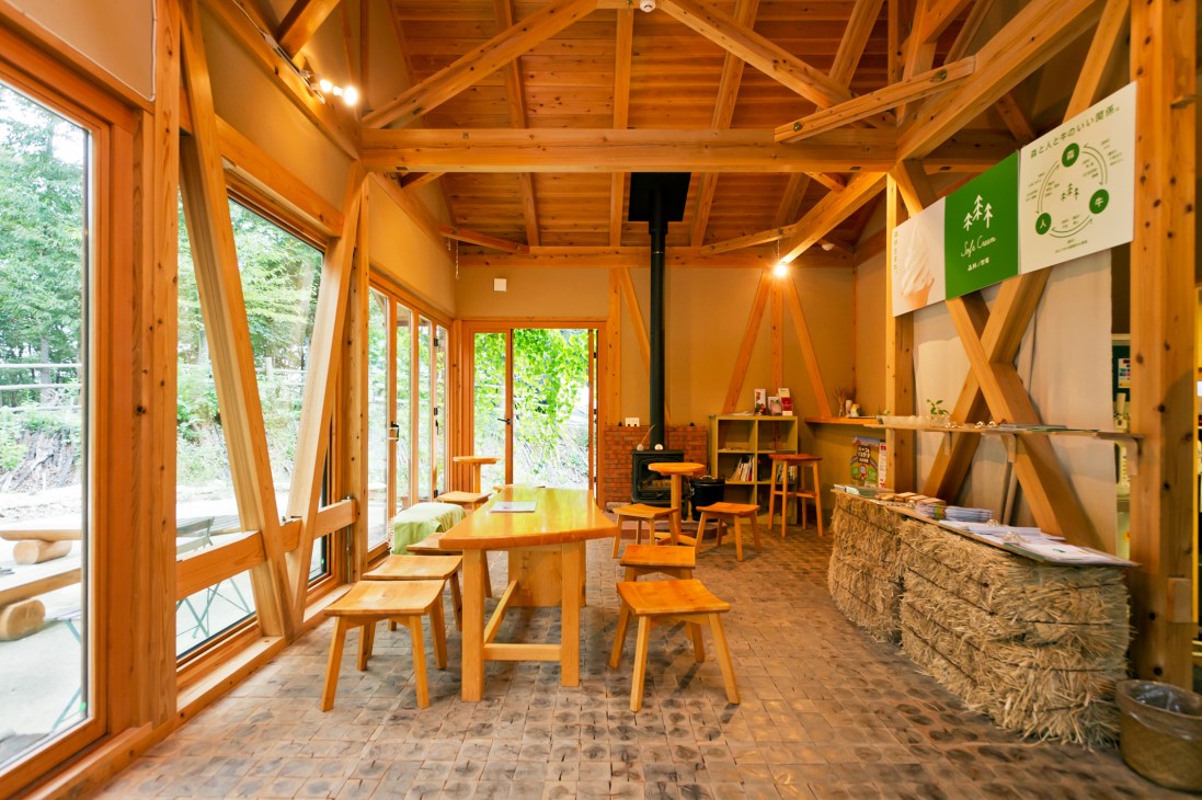 Interior of a wooden café building with natural light, tables, chairs, and a wood-burning stove at Yuimarl Nasu.