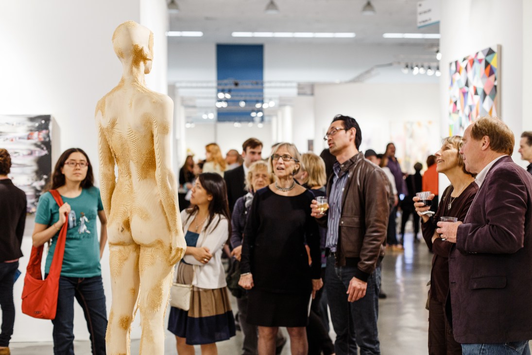 Gallery visitors observe a tall wooden sculpture of a nude female figure at an art fair.