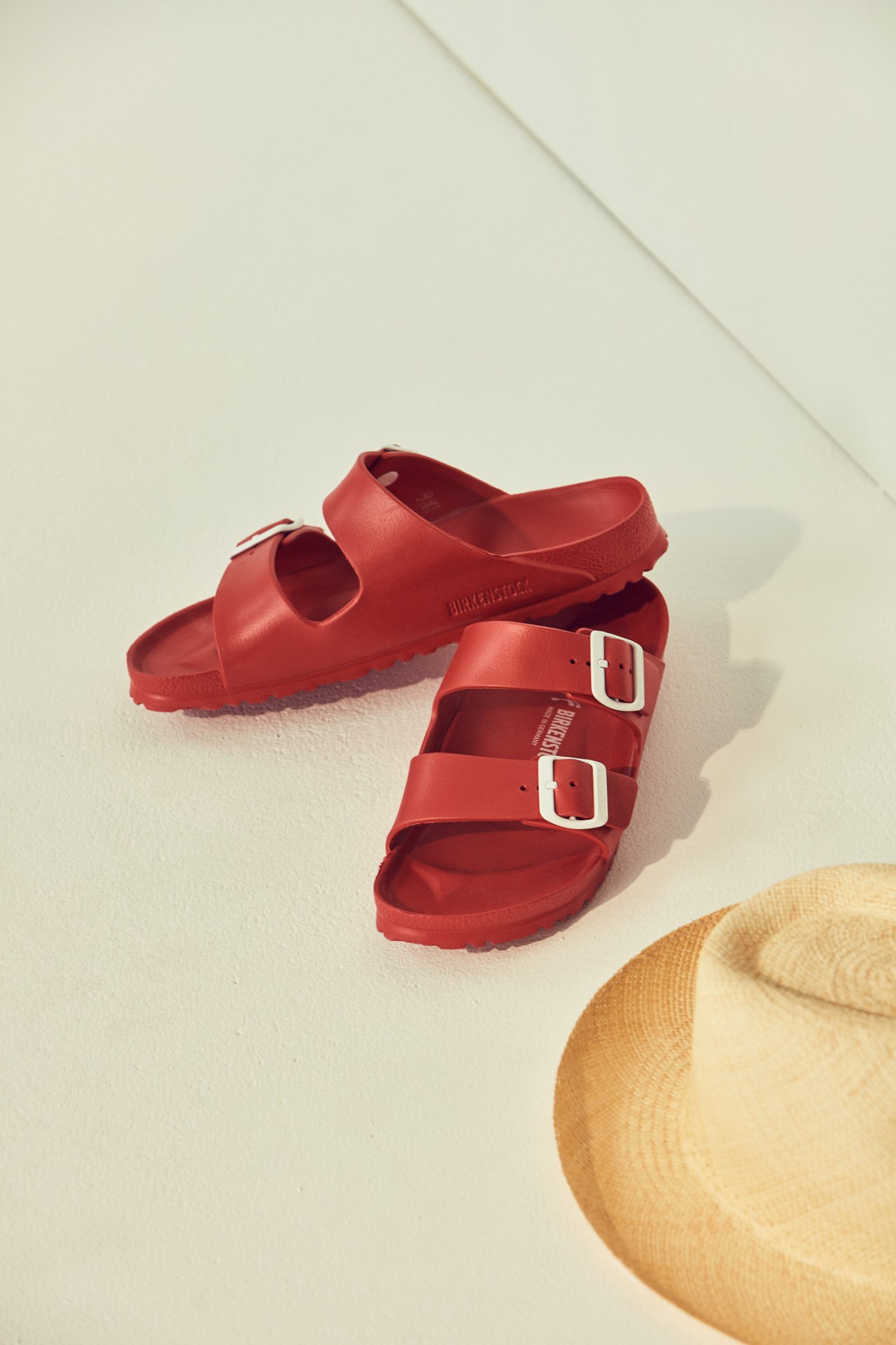 Red Birkenstock sandals with white buckle straps on a white surface next to a straw hat.
