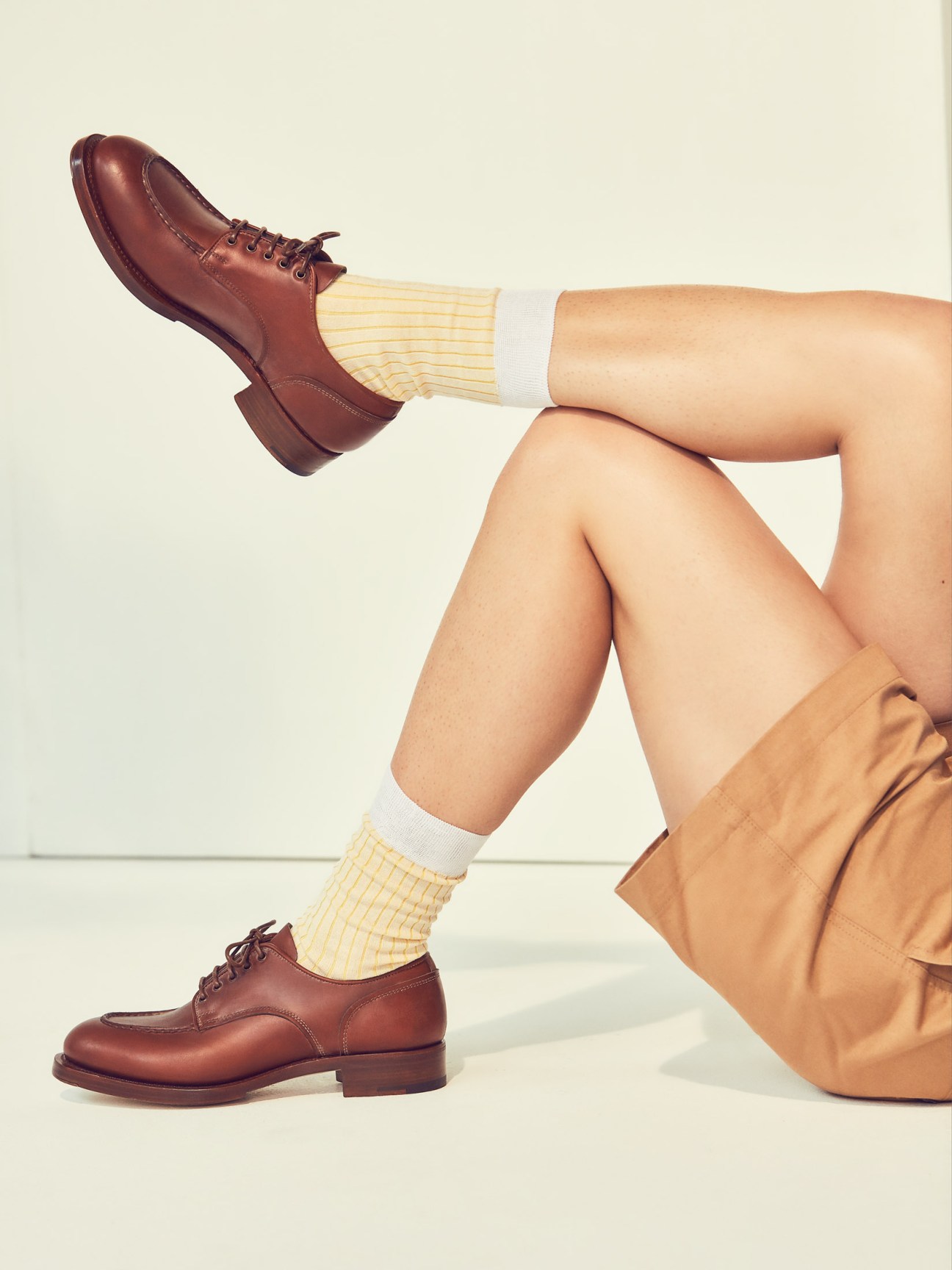 Model's legs wearing yellow socks, brown leather shoes by Santoni, and tan shorts by Maison Kitsuné.