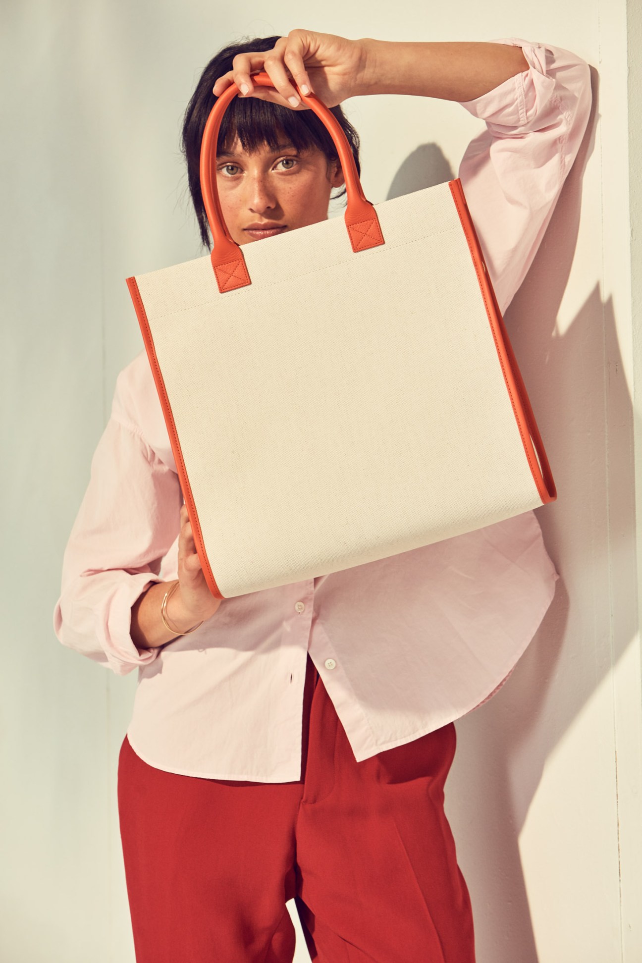 Woman holding a cream and coral tote bag by L/Uniform, wearing a pink shirt and red trousers.