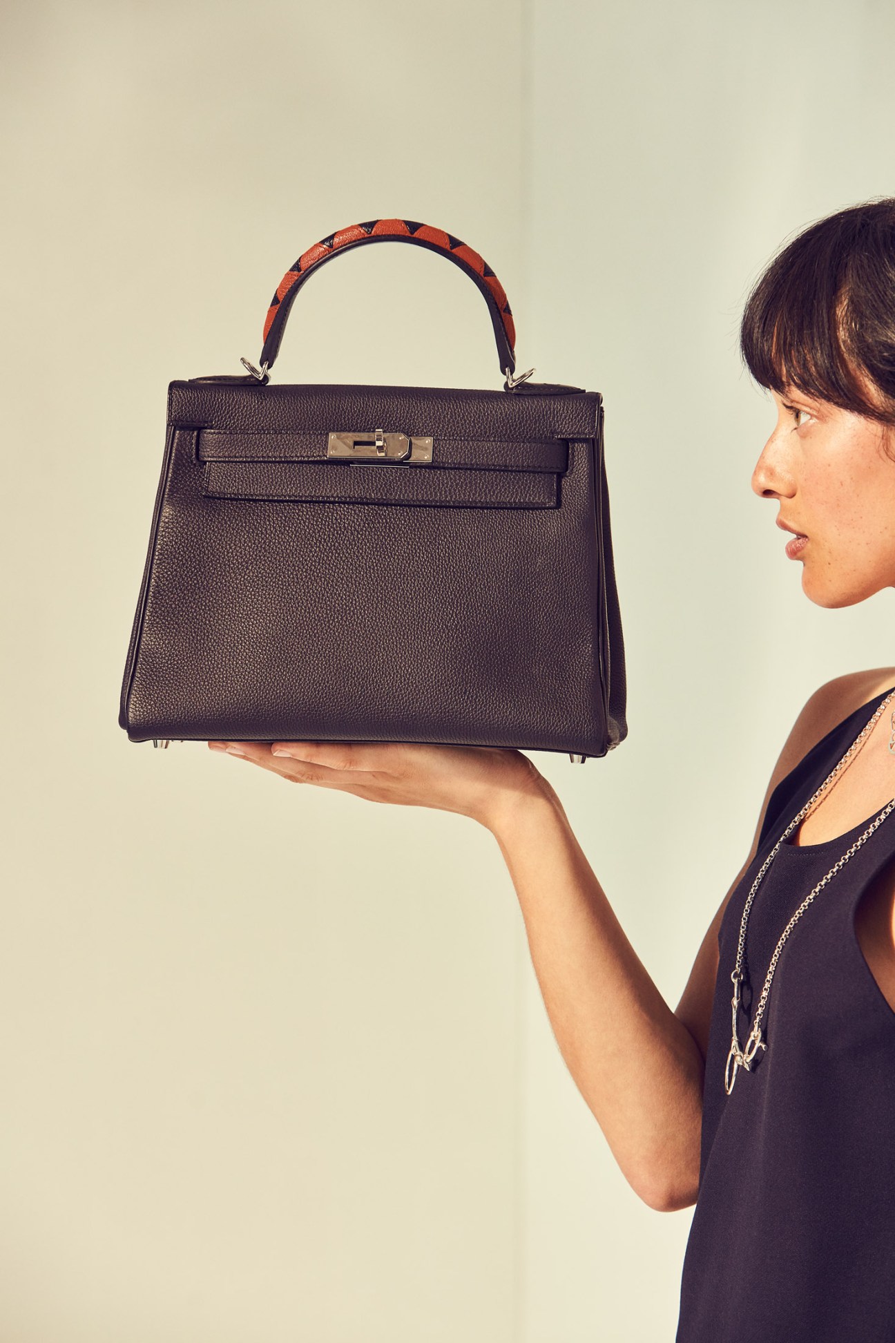 Woman holding a black leather Hermès bag with red tortoiseshell handle against neutral background.