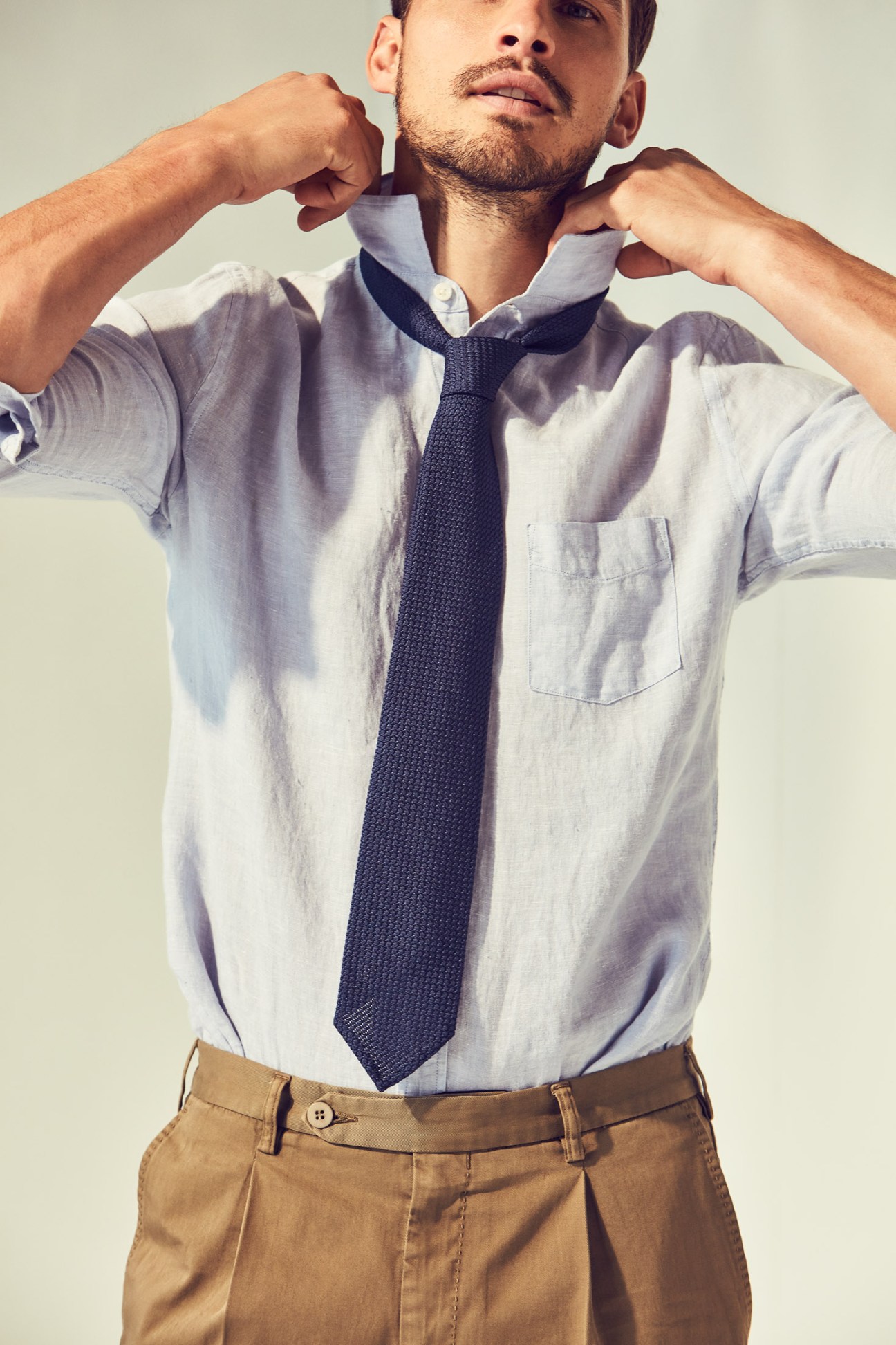 Man wearing a light blue shirt, dark tie, and khaki shorts against a neutral background.