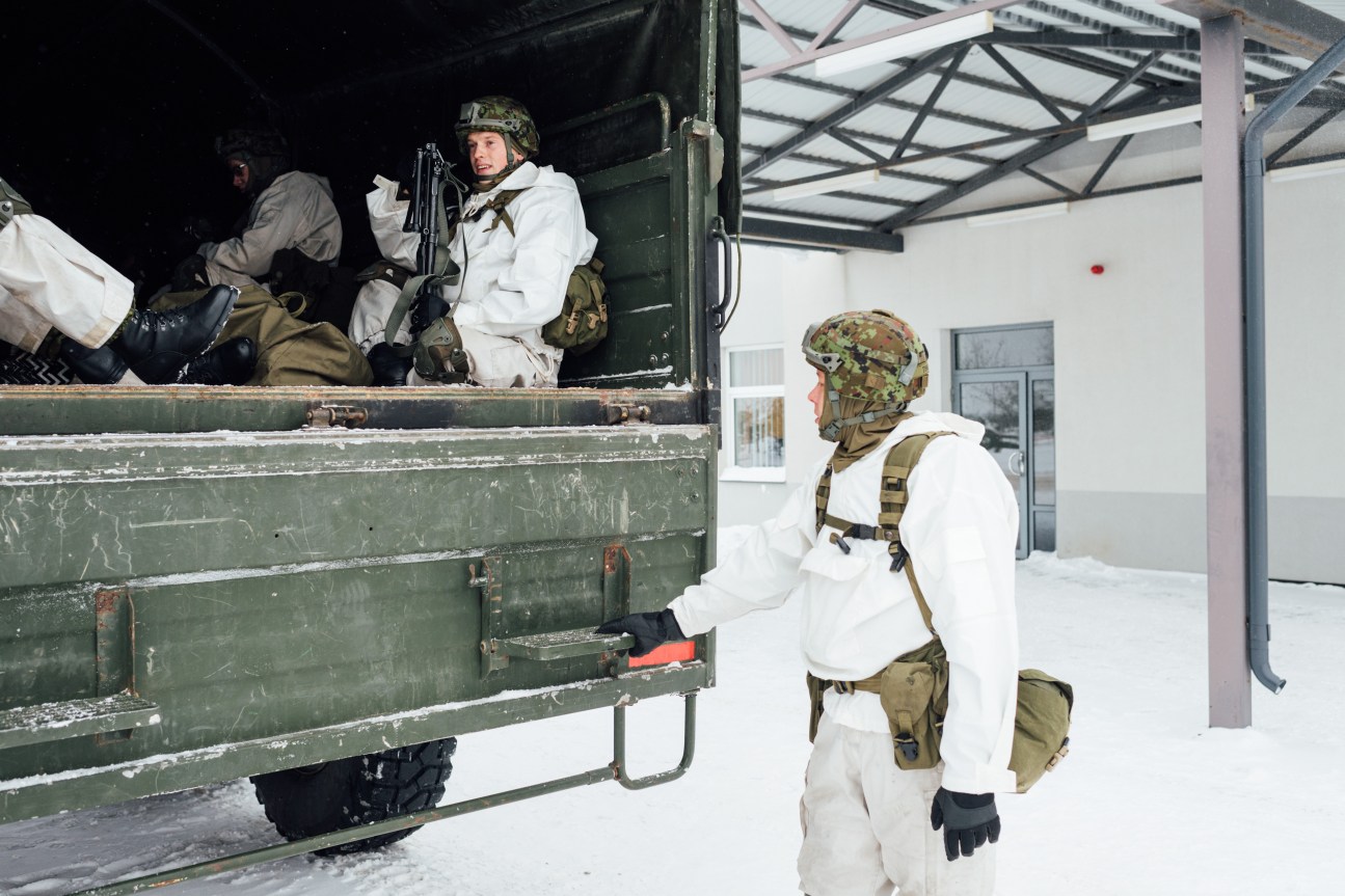 Soldiers in white winter camouflage loading onto a green military truck at a snowy army base.