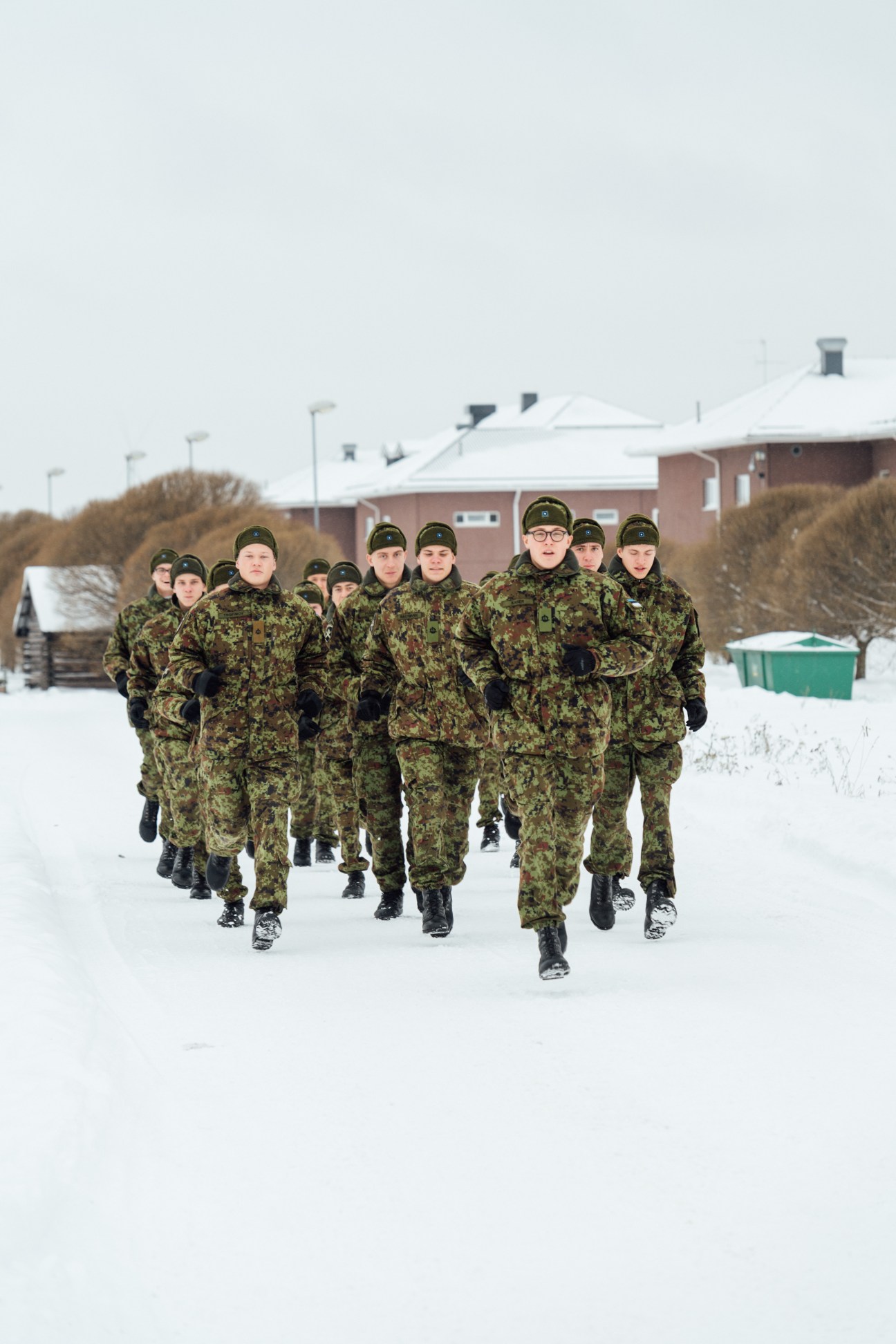 Estonian soldiers in camouflage uniforms jogging through snow during a military drill at a base.