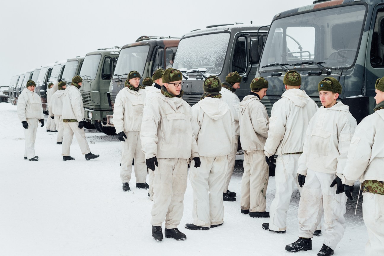 Estonian soldiers in white winter uniforms stand in front of military convoy trucks covered in snow.
