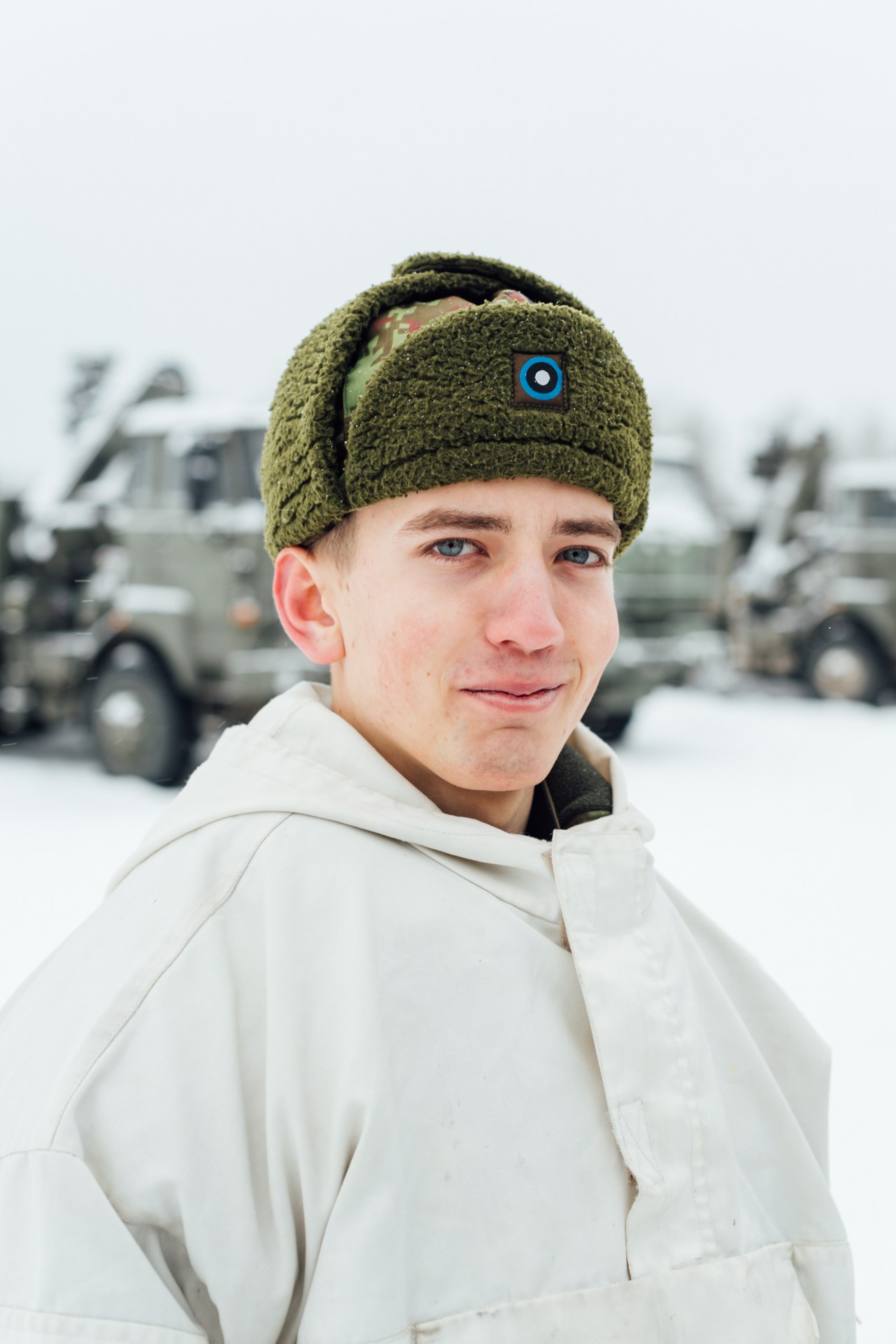 Young Estonian soldier in olive green knit hat and white jacket at military base with vehicles blurred in background.