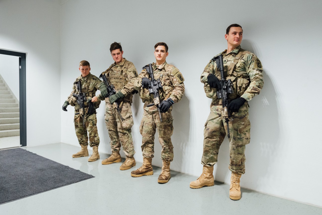 Four US soldiers in desert camouflage uniforms and combat boots stand in a line holding rifles indoors.