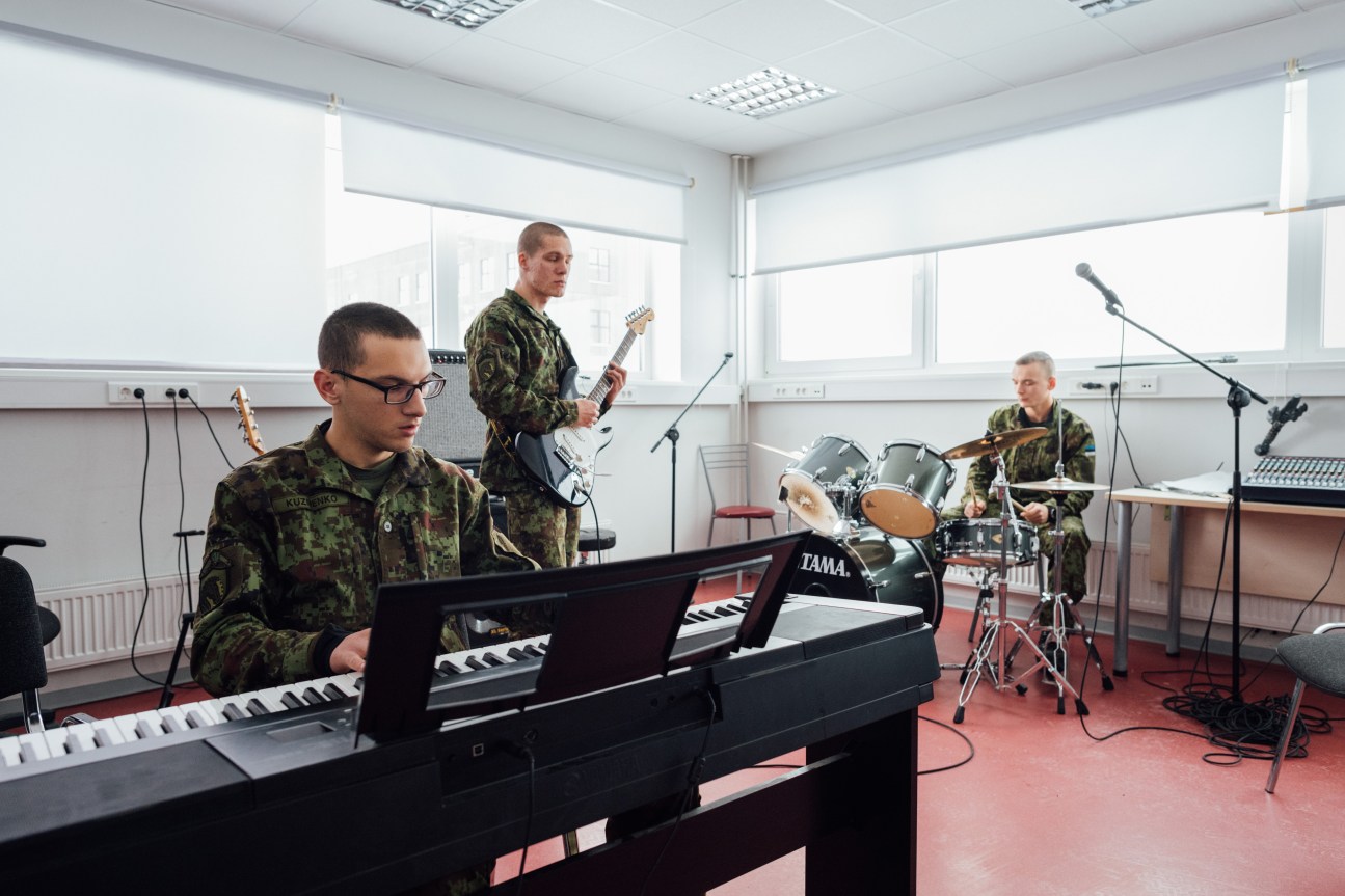Estonian soldiers in camouflage uniforms performing music together in a bright room with a keyboard, guitar, drums and microphones.
