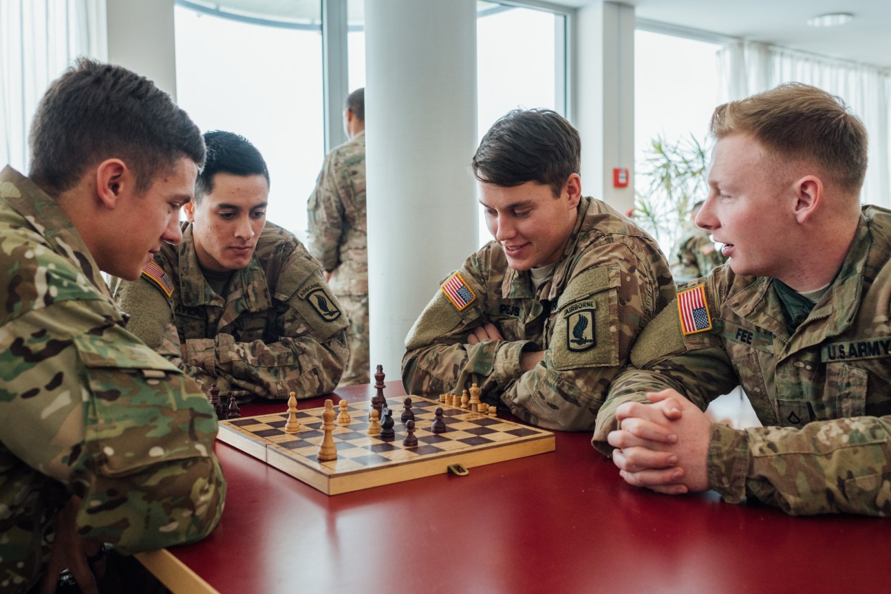 US Army soldiers in camouflage uniforms play chess indoors at a military base.