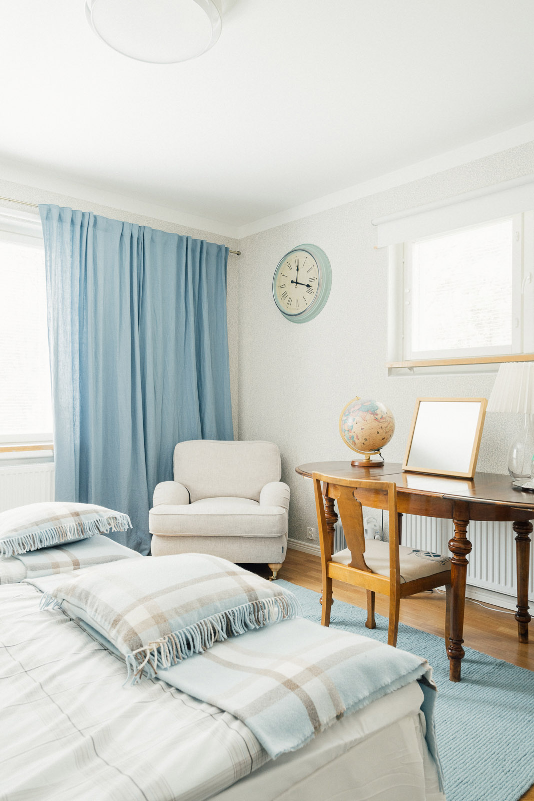 A bright, minimalist bedroom with a cream armchair, wooden desk, blue curtains, and striped bedding in soft tones.