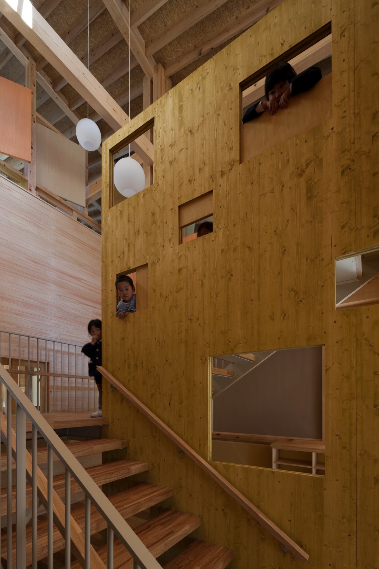 Children peek through cutout windows in wooden walls of a Japanese kindergarten interior.