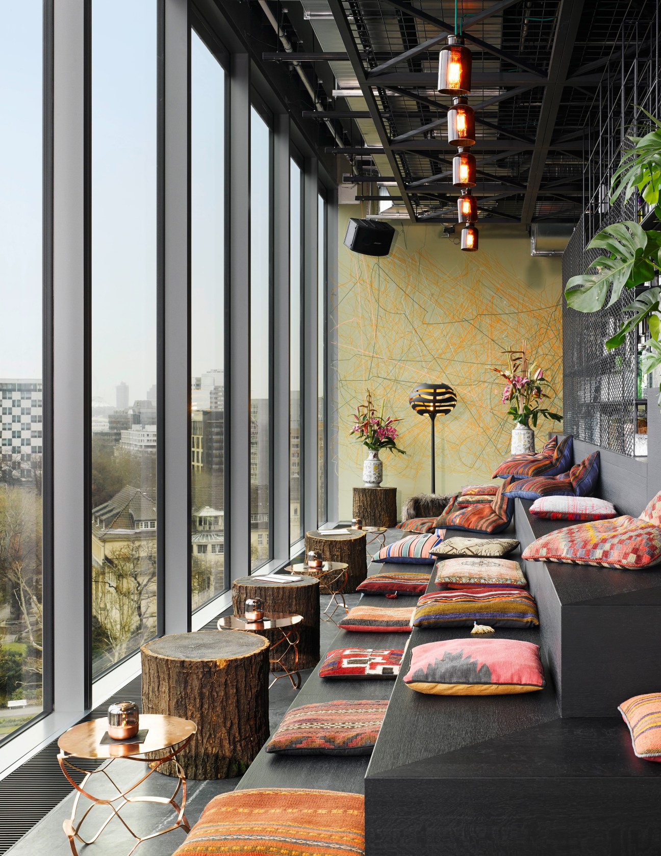 Modern rooftop lounge with colorful patterned cushions, tree-stump tables, hanging lanterns and city views.