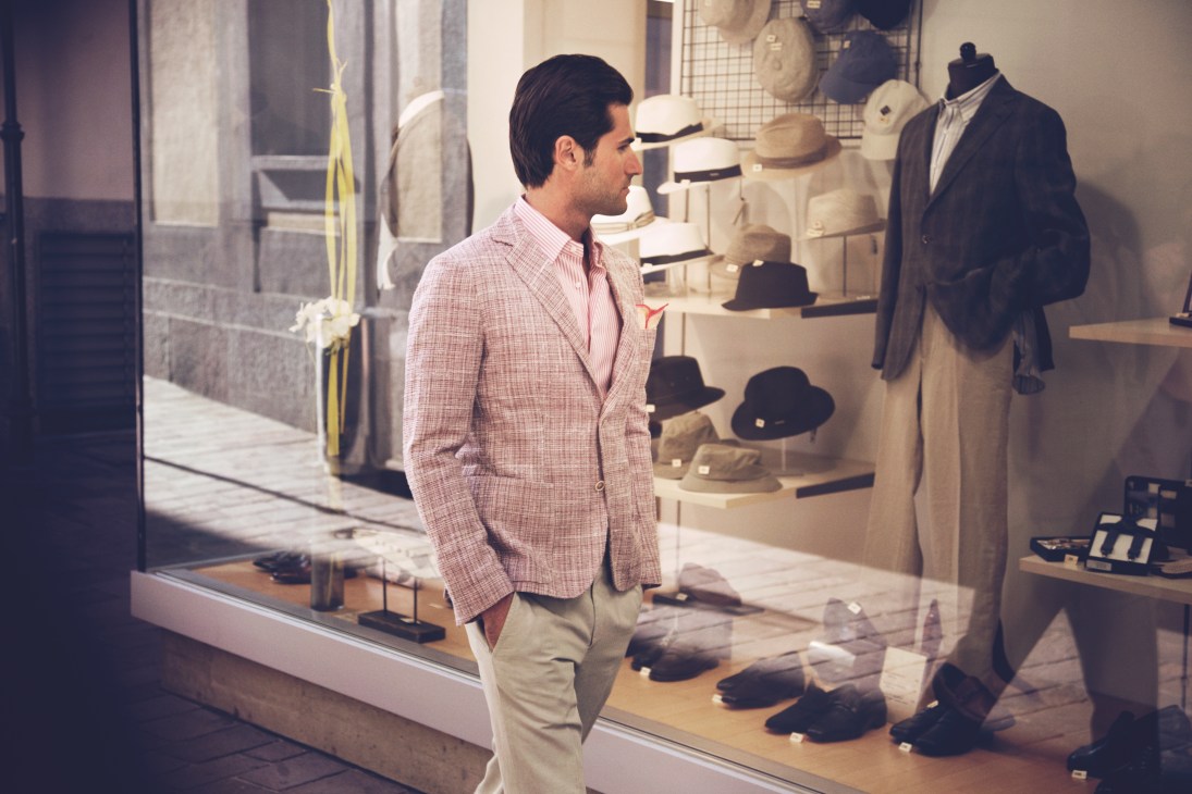 Man in pink jacket and beige trousers looking at hat display in shop window in Lugano.