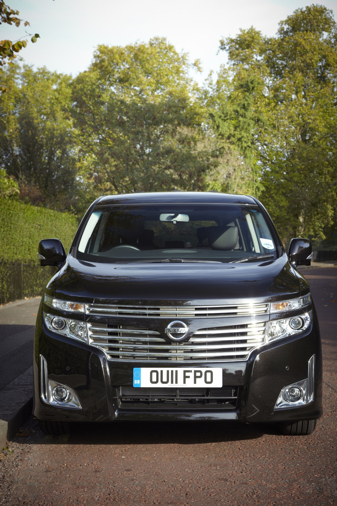Front view of a black Nissan Elgrand Highway Star minivan parked on a driveway with green trees in the background.