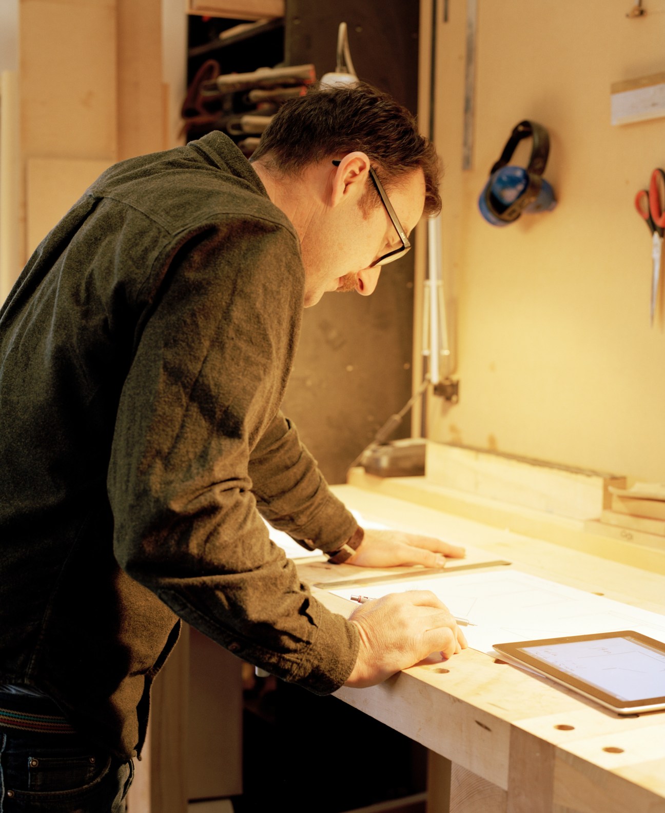 Craftsman in workshop wearing glasses carefully measures and marks wood for furniture making.