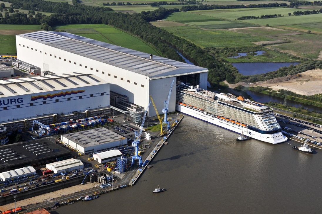 Large cruise ship docked at Meyer Werft shipyard in Germany with industrial facilities and green countryside visible.