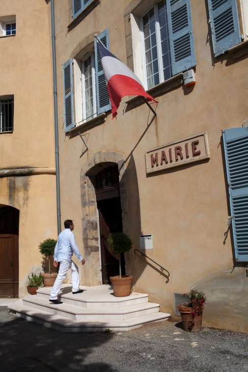 Man in white clothing stands before a stone mairie building with French flag and blue shutters in Tourtour.