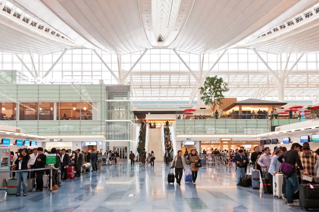 Bright, modern airport terminal interior with high ceilings, natural light, and travelers with luggage.
