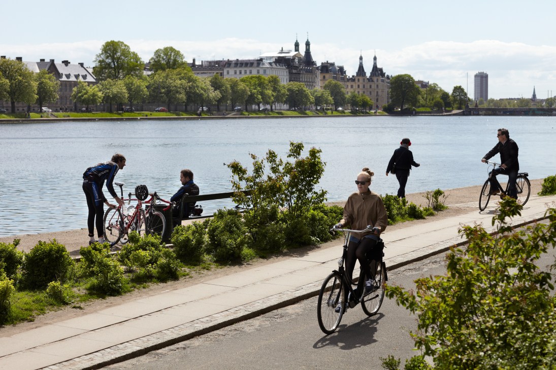 Cyclists and pedestrians enjoy a waterfront path along a Copenhagen canal with historic buildings in the background.