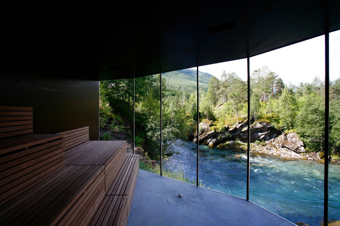 Glass-walled sauna with wooden bench overlooking a river and forested mountains through large windows.