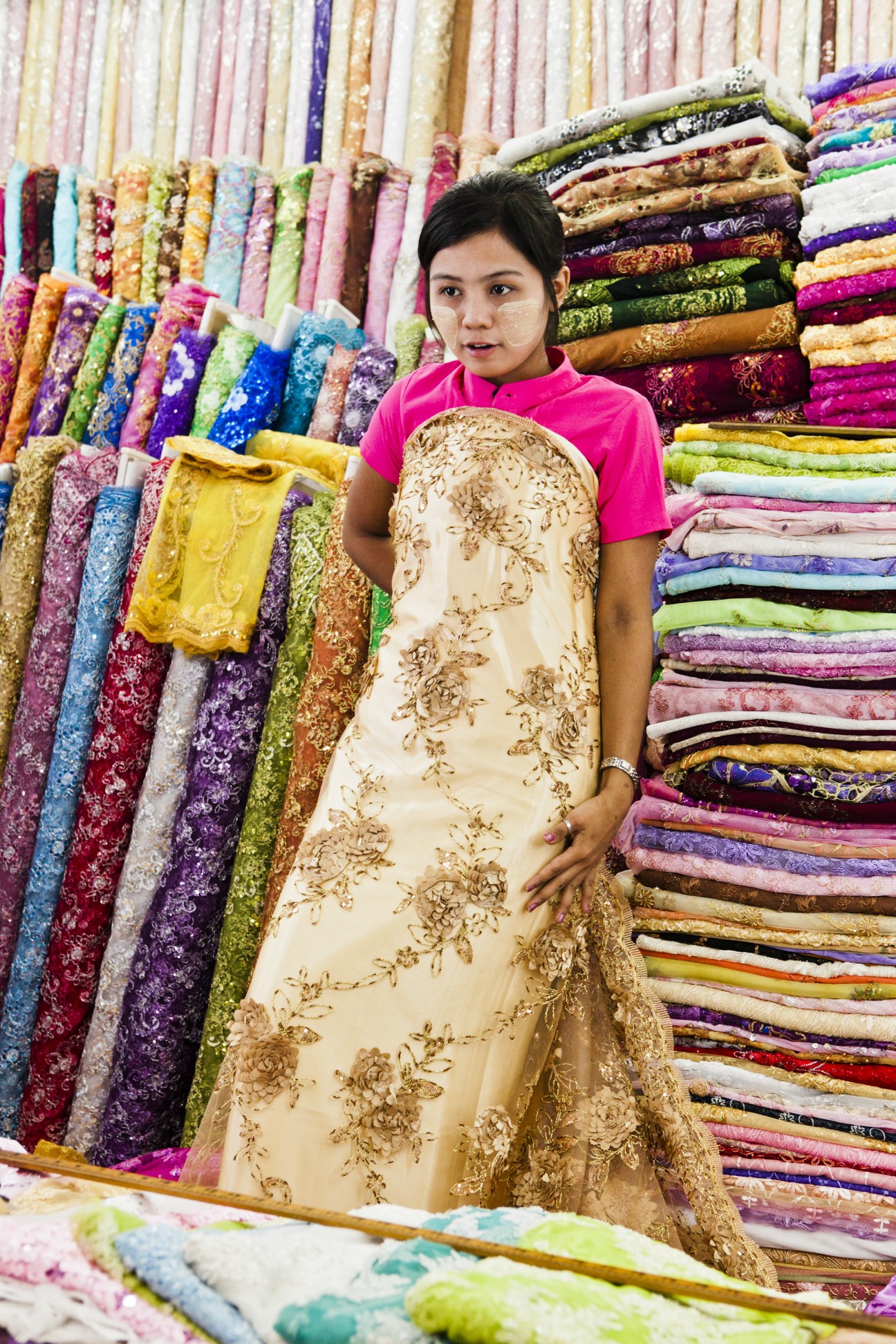 A vendor in a pink shirt models an ornate cream and gold patterned fabric at Scott Market in Rangoon, surrounded by colorful stacked textiles.