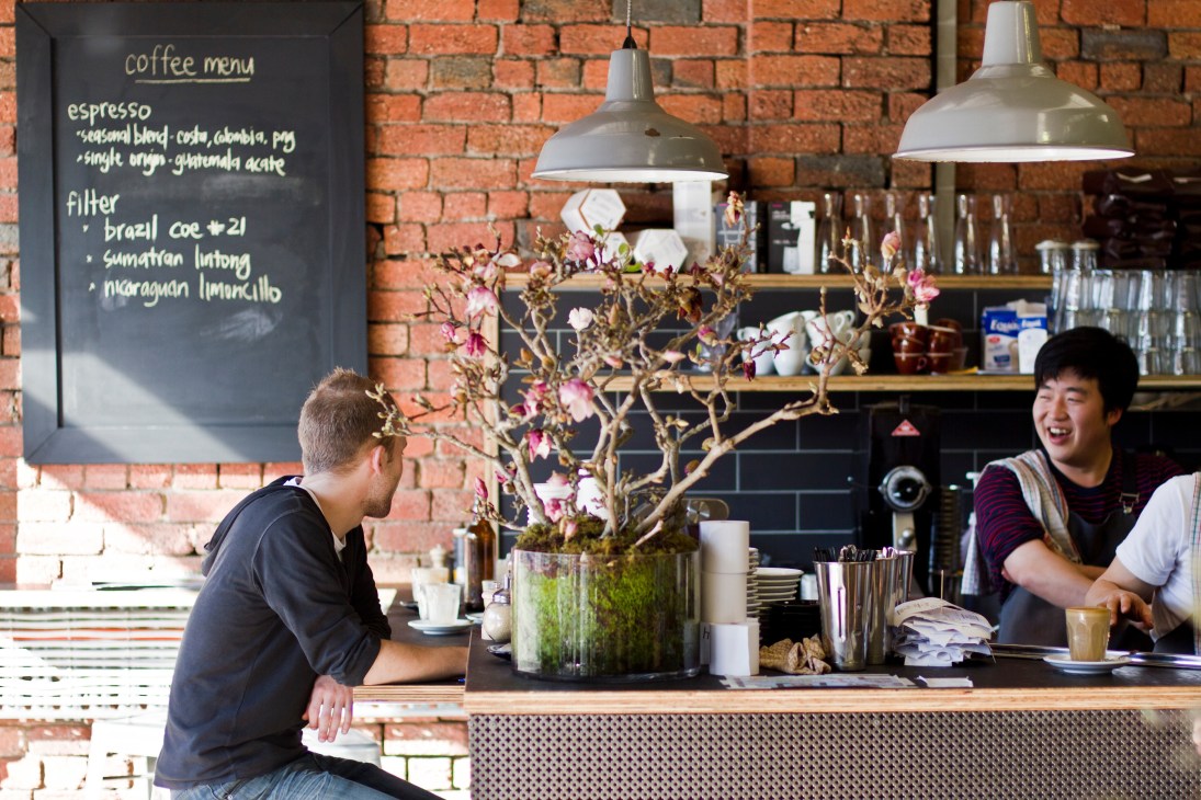 Customer at Hobba café counter with barista, exposed brick walls and coffee menu board visible.