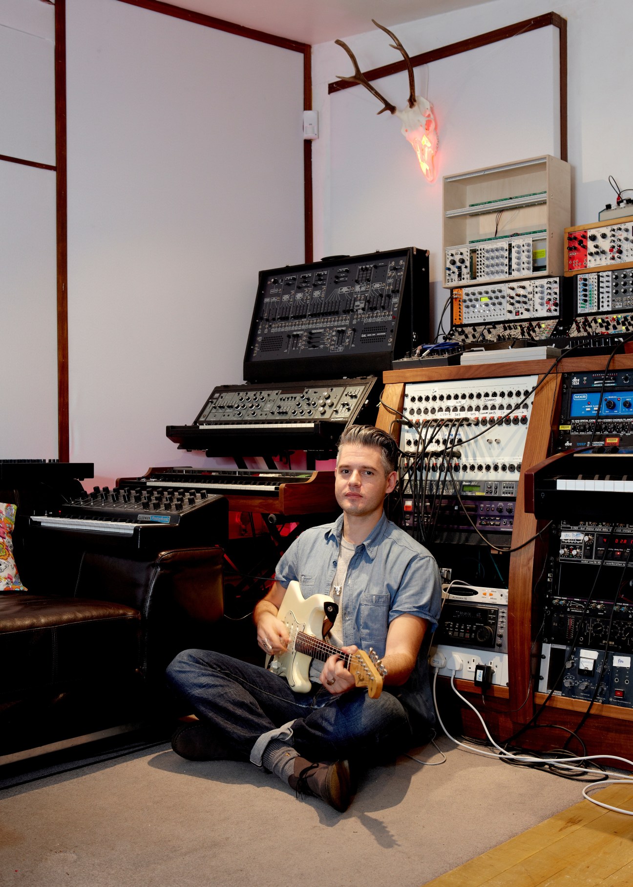 Music producer Paul Epworth sits with a guitar in his studio surrounded by synthesizers and electronic equipment.
