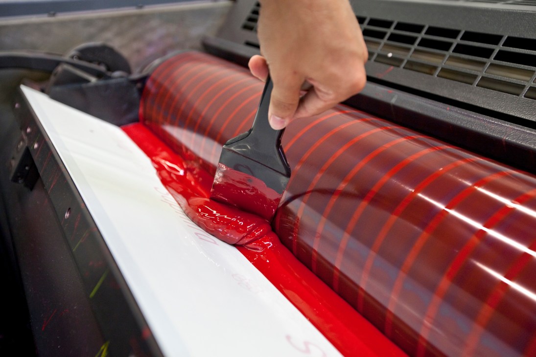 Technician loading red ink onto a Heidelberg printing press roller.