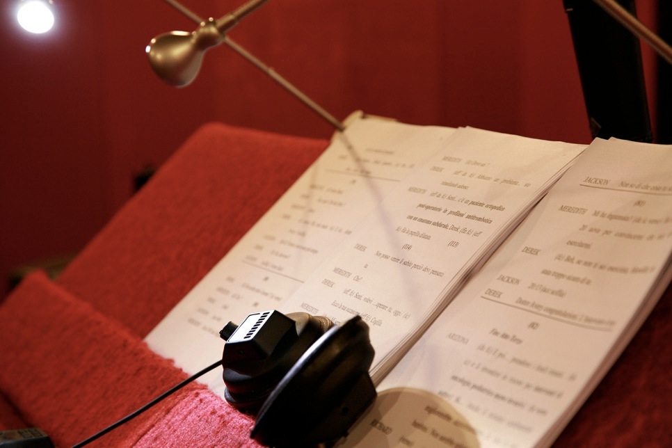 A black voice actor's earpiece lies on an open script under a desk lamp in a red-walled dubbing studio.