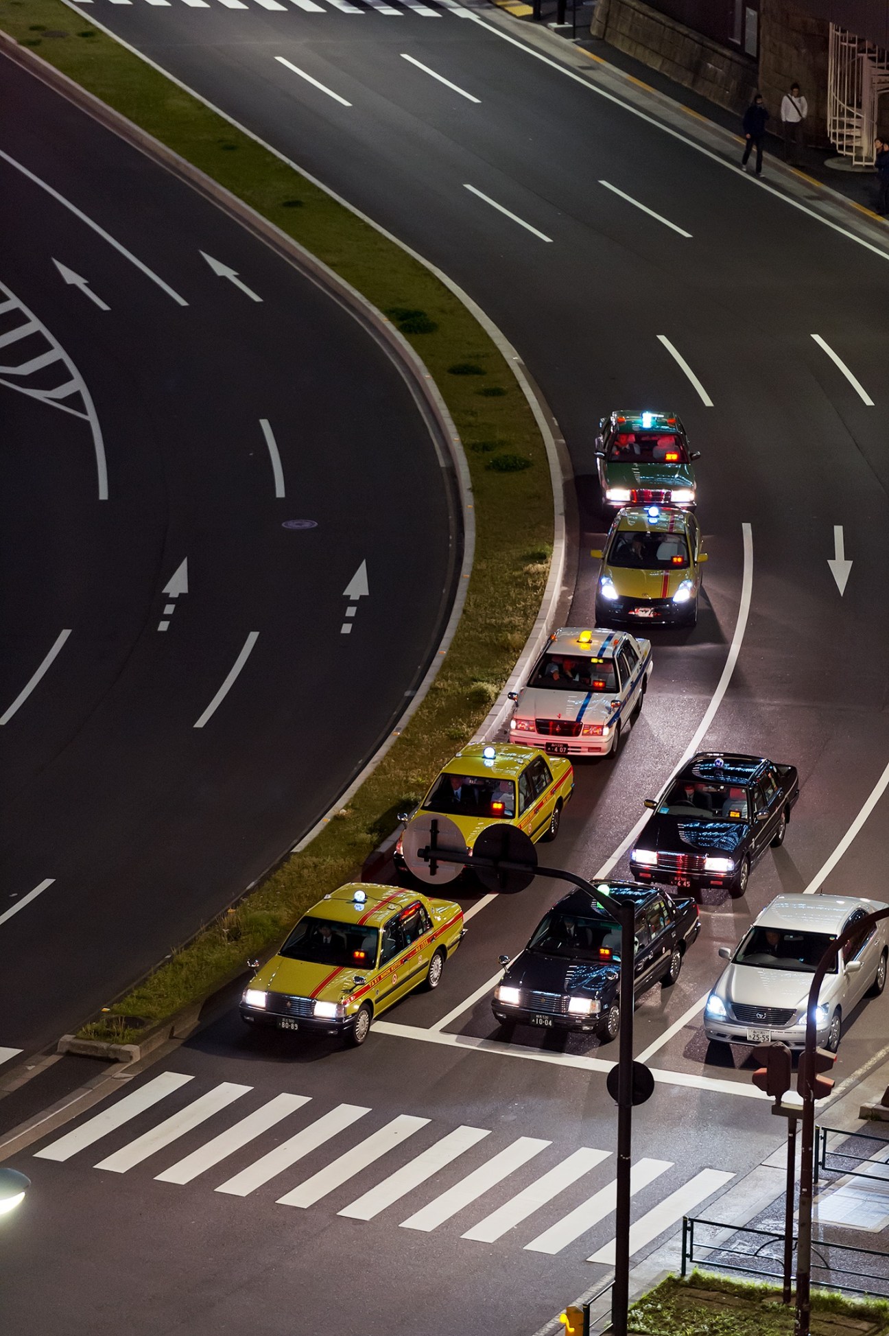Tokyo taxis lined up on a curved road at night, displaying yellow, black and white vehicles with illuminated headlights and rooftop lights.