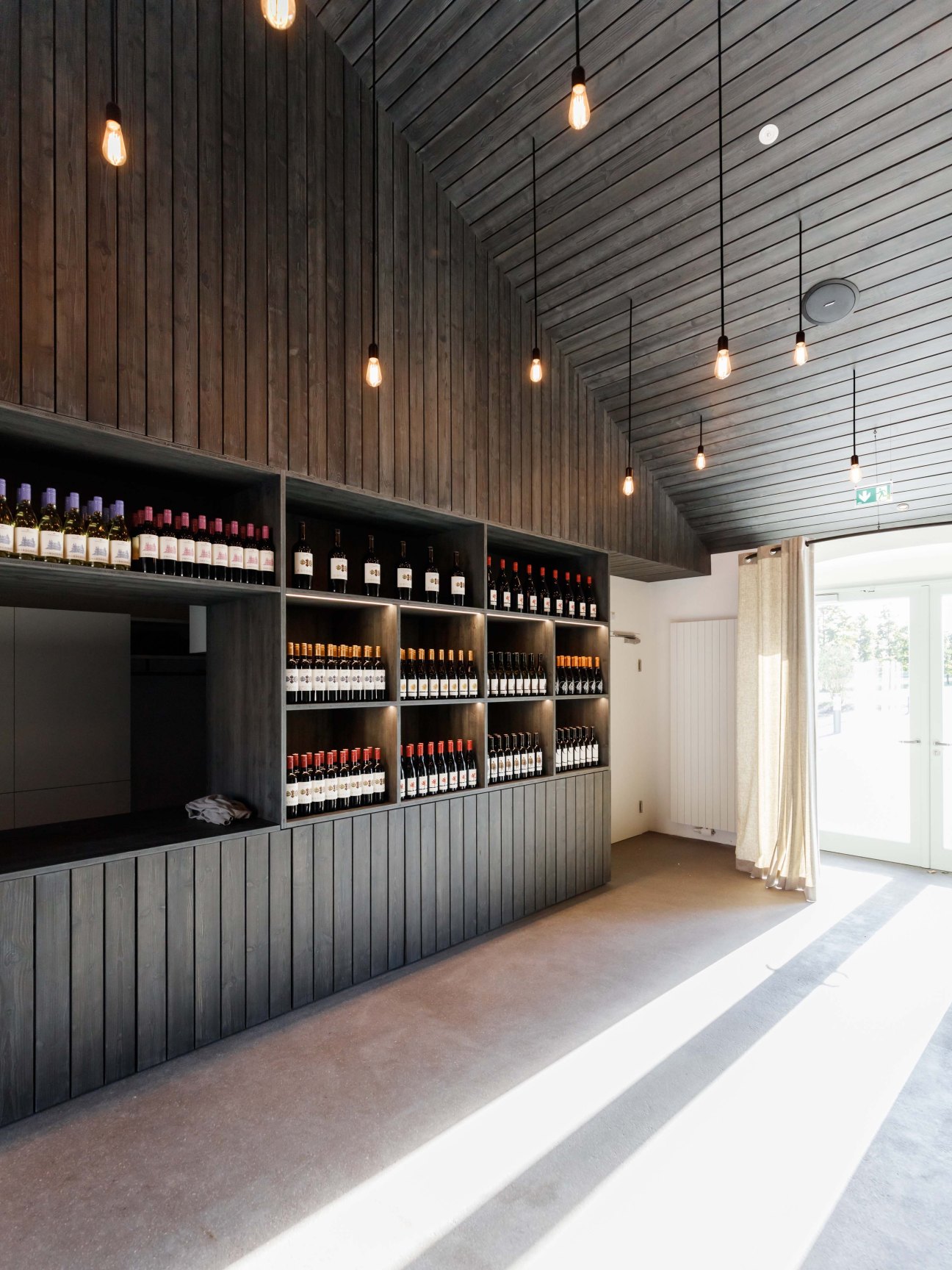 Modern bar interior with dark wood slat ceiling, pendant lights, and wine bottles displayed on wall shelving.
