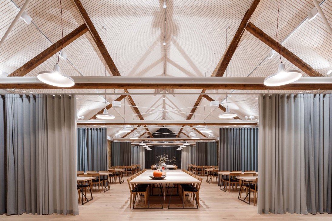 Interior of a refitted farmhouse with exposed wooden beams, pendant lights, and wooden dining tables arranged in a spacious room with grey curtains.