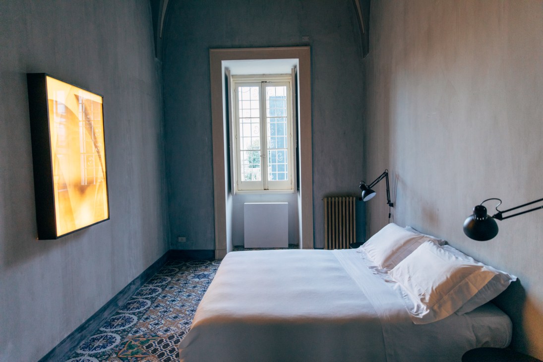 Minimalist bedroom with white bed, illuminated yellow artwork, black desk lamp, and patterned mosaic floor in Palazzo Daniele hotel.