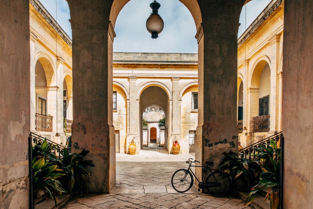 Neoclassical courtyard of Palazzo Daniele with arched columns, terracotta floor, hanging lamp, and potted plants.