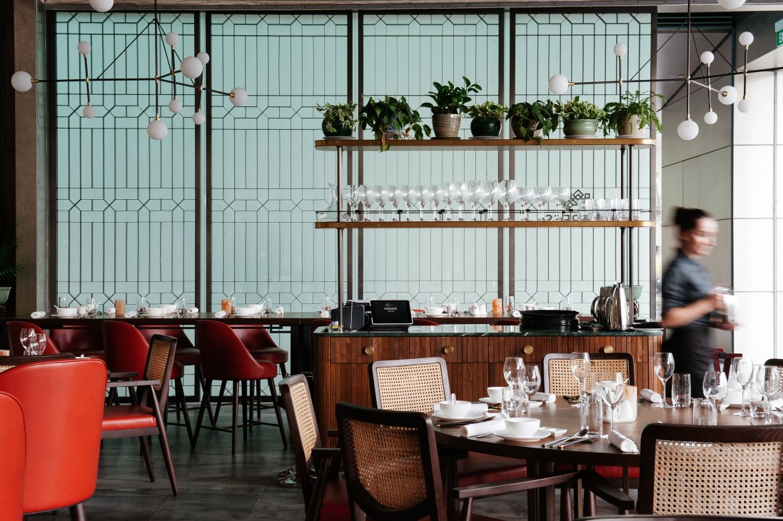 Modern restaurant interior with green tile wall, cane-backed chairs, and suspended shelving with plants