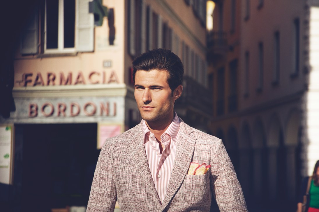 Man in light pink jacket and striped shirt standing on an Italian street outside a pharmacy.