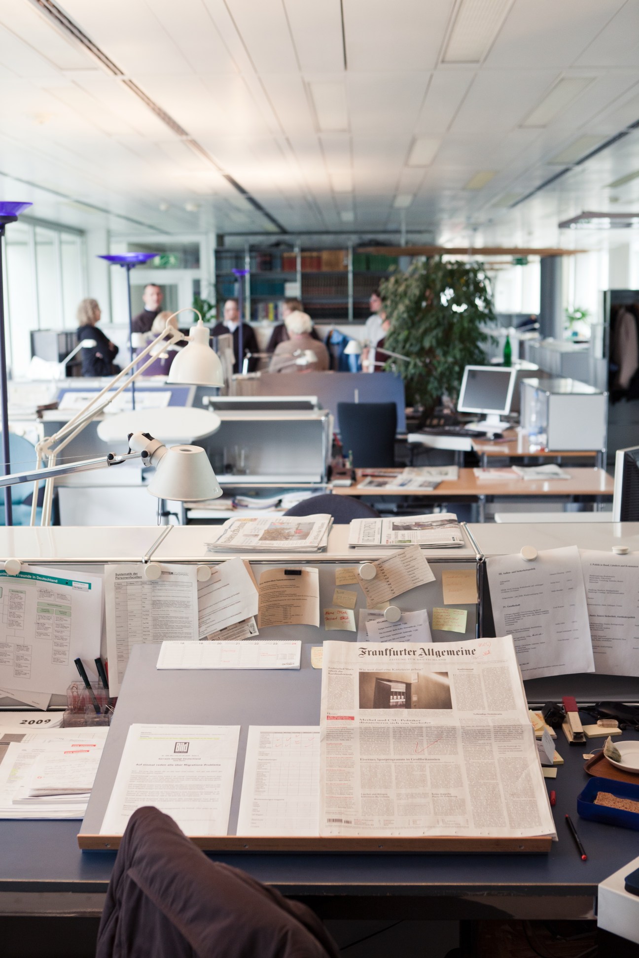 An organized newsroom desk with newspapers and documents spread out, with blurred staff members working in the background at Der Spiegel's office.