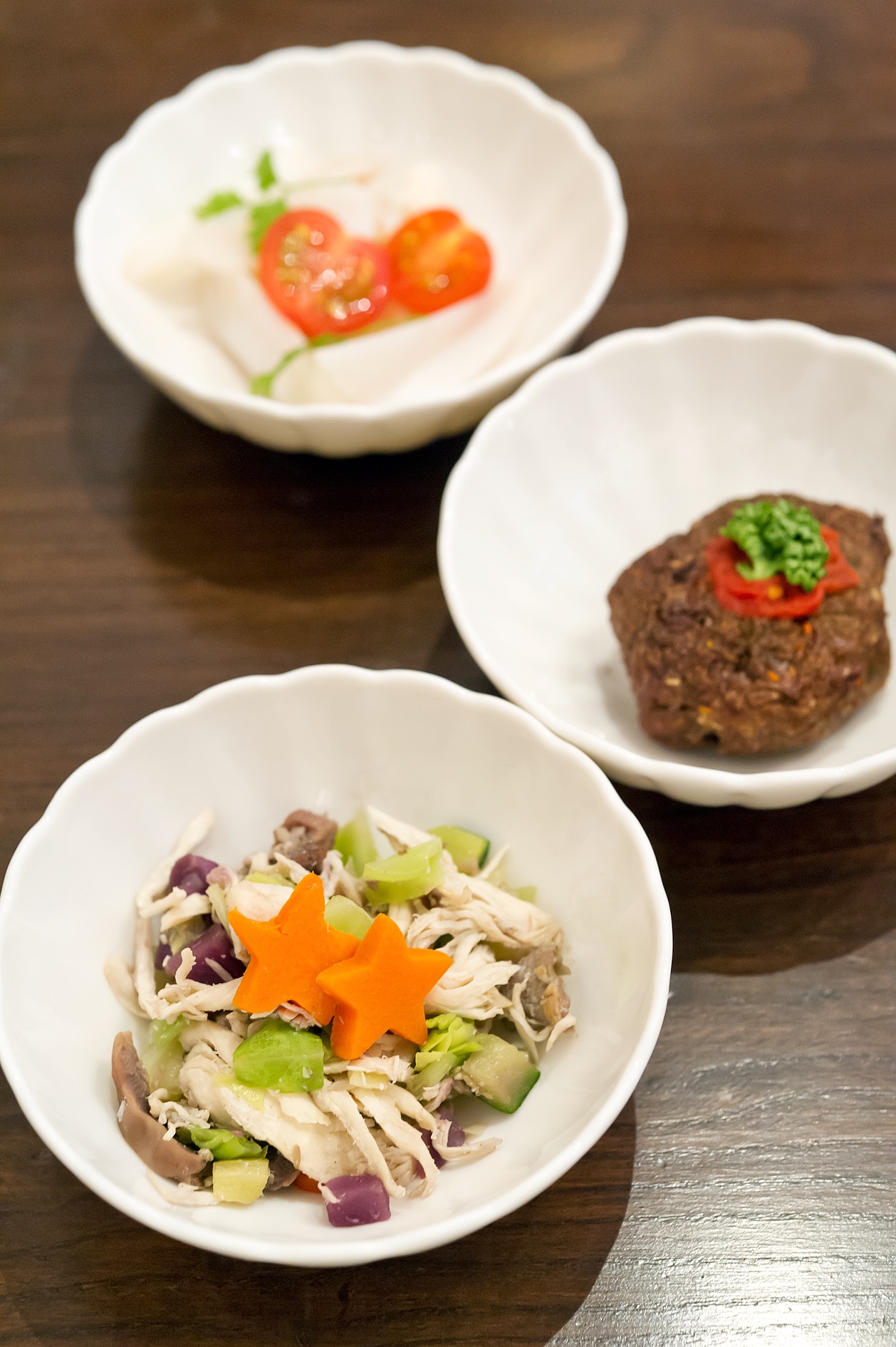 Three white bowls of gourmet dog food: chicken salad with vegetables, horse meatballs with red pepper, and a white creamy dish with tomato garnish.