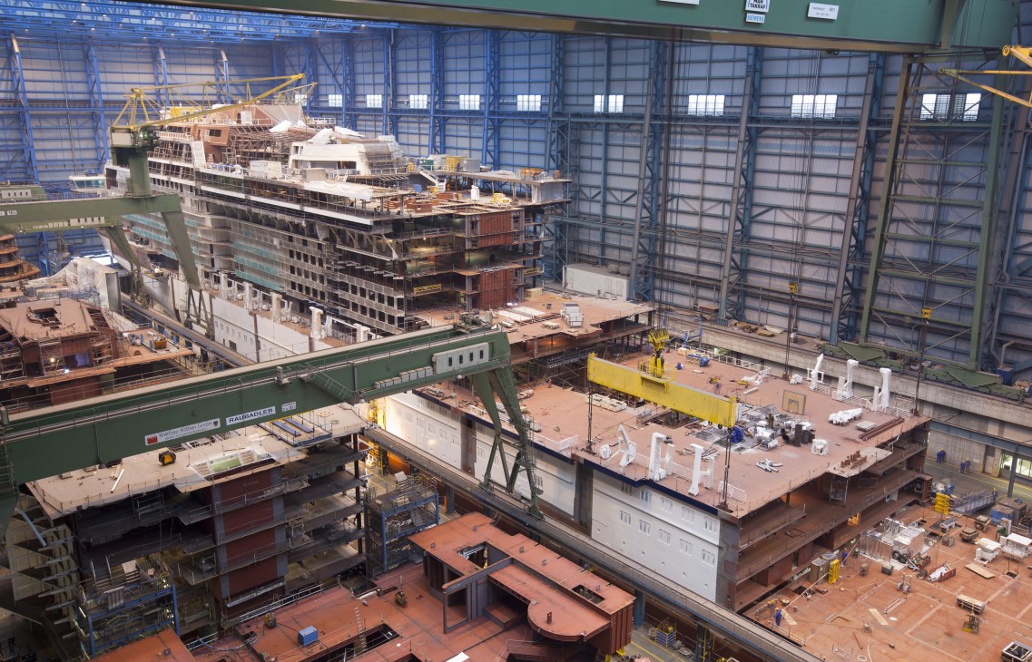 Large cruise ship under construction inside an industrial shipyard with metal framework and crane equipment.