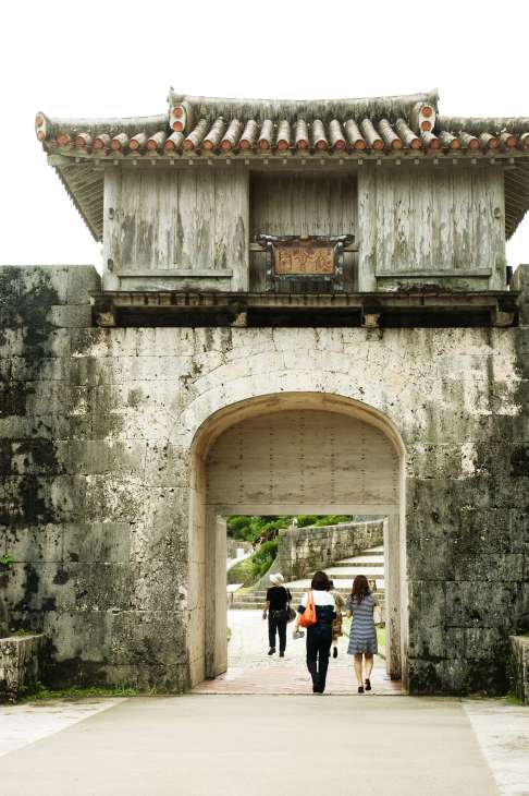 Traditional Okinawan gate with curved tile roof and weathered wooden structure, visitors passing through stone archway.