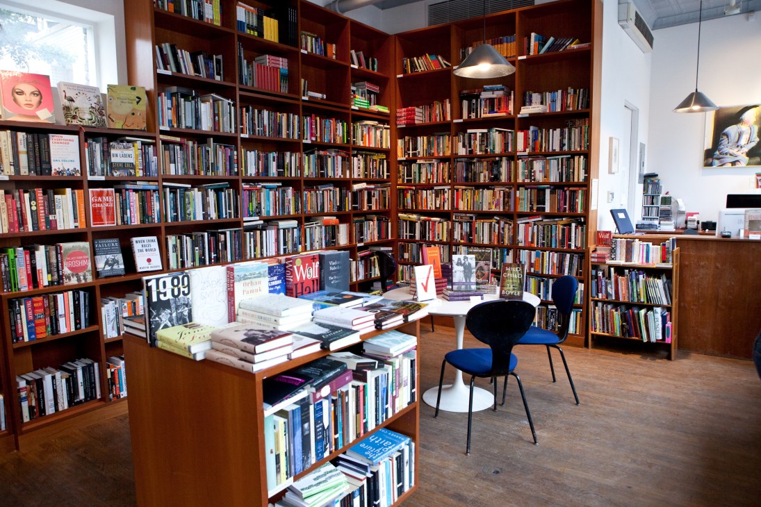 Interior of 192 Books bookstore with floor-to-ceiling wooden shelves filled with books, a central display table, and blue chairs.