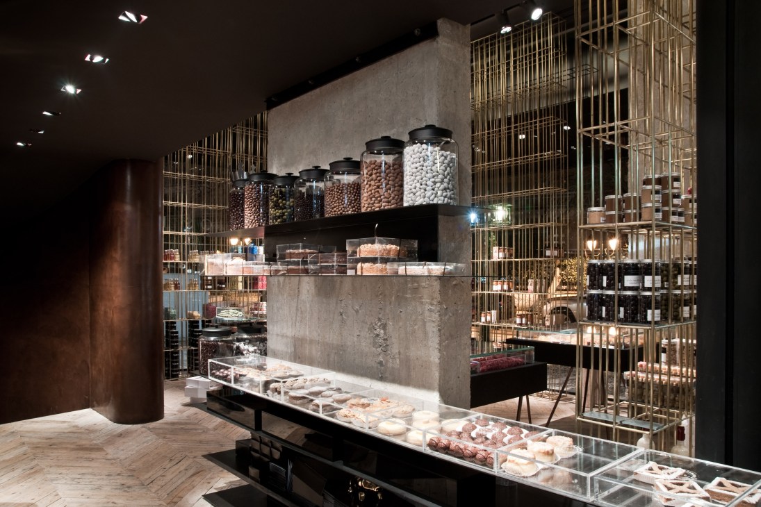 Modern pastry shop interior with concrete walls, brass caging, display cases of baked goods and jars of ingredients on shelves.